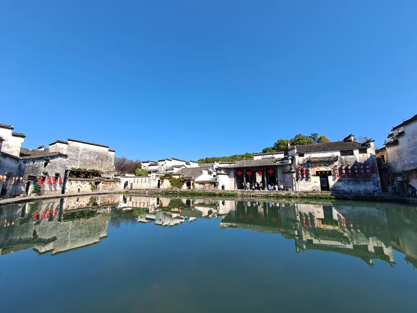 蓝天下月沼全景，环池白墙黛瓦民居、红灯笼与岸边人群倒映在水中